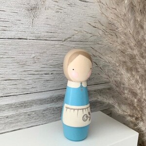 May include: A wooden doll with a light blue dress and a white apron. The doll has brown hair and a painted face with pink cheeks.