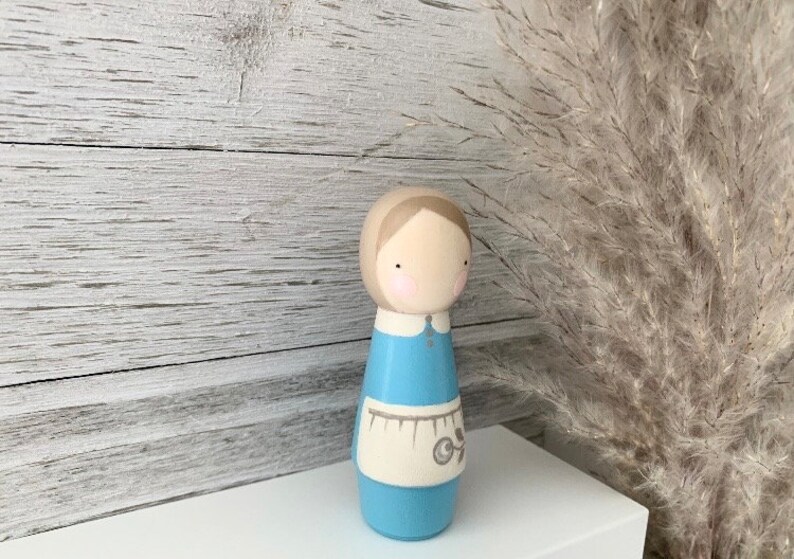 May include: A wooden doll with a light blue dress and a white apron. The doll has brown hair and a painted face with pink cheeks.