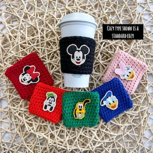 May include: A set of six crocheted coffee cup cozies featuring different Disney characters. The cozies are in red, green, blue, pink, and black. The characters include Mickey Mouse, Minnie Mouse, Donald Duck, Goofy, and Pluto.
