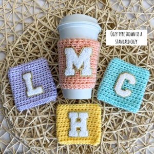 May include: A peach-colored crochet cup sleeve on a white cup, surrounded by three square crochet coasters in lavender, turquoise, and yellow. Each item features a white varsity-style letter with a gold glitter border. Text reads: COZY TYPE SHOWN IS A STANDARD COZY.