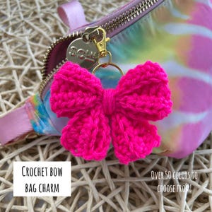May include: A bright hot pink crochet bow bag charm attached to a gold-toned metal lobster clasp and ring. The charm is clipped onto a tie-dye fanny pack featuring pastel pink, yellow, and blue colors with a gold zipper and a circular metal tag.