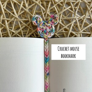 May include: A colorful crocheted mouse bookmark with a long, braided tail. The bookmark is on a white book with the text "CROCHET MOUSE BOOKMARK" on a white label.