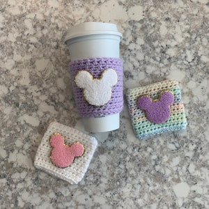 May include: A white reusable coffee cup with a lavender crochet sleeve featuring a white mouse-ear patch with gold trim, alongside a white crochet coaster with a pink mouse-ear patch and a pastel variegated crochet coaster with a purple mouse-ear patch.