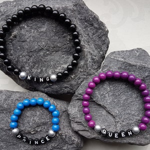 Könnte beinhalten: Drei Perlenarmbänder mit silbernen Perlen und schwarzen Buchstaben, die "KING", "QUEEN" und "PRINCE" buchstabieren. Die Armbänder liegen auf einem grauen Steinuntergrund.