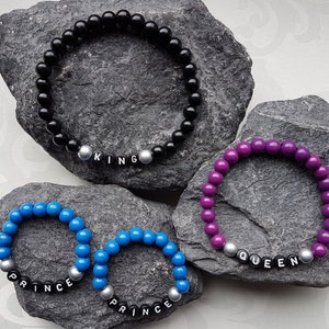 Könnte beinhalten: Vier Perlenarmbänder mit silbernen Buchstabenperlen, die "KING", "QUEEN" und "PRINCE" buchstabieren. Die Armbänder sind schwarz, lila und blau.