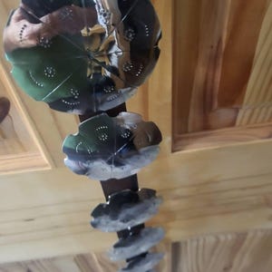 May include: A decorative hanging mobile featuring a series of metallic, flower-shaped elements. The mobile has a dark brown central rod with several tiers of silver and green flower-like shapes, and a series of stacked, gray, stone-like elements.