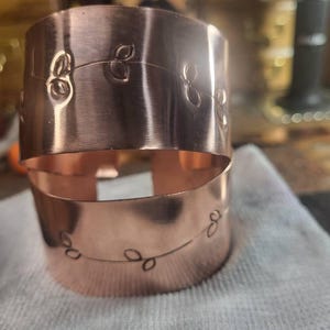 May include: Two copper cuff bracelets stacked. Each bracelet has an etched design of a vine with leaves. The bracelets are a shiny, warm copper color. The background is blurred, suggesting a workshop setting.