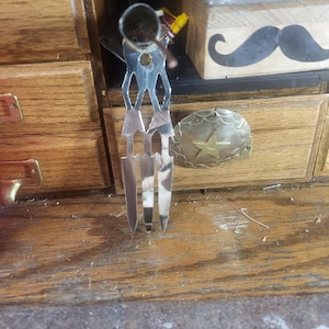 May include: A silver decorative fork with a distinctive design. The fork has a handle shaped like a person with arms and legs, and three tines. Next to it is a brass-coloured, star-shaped decorative piece.