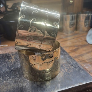 以下が含まれることがあります： 金色の金属製ナプキンリング2つ。彫刻された花柄模様が施されています。リングは重ねられており、複雑なデザインを示しています。背景はぼやけており、ワークショップの環境を示唆しています。