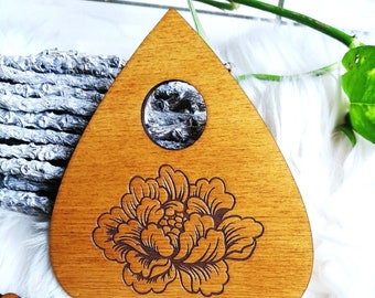 May include: A wooden Ouija board with a floral design and a hole in the shape of a crescent moon. The board is brown and has a light wood grain.