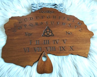 May include: A wooden Ouija board with engraved letters, numbers, and the words "Goodbye", "Yes", and "No". The board has a dark brown finish and includes a planchette. The background is a fluffy white surface.