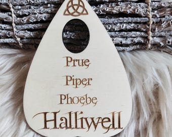 May include: A light-colored wooden planchette with the names Prue, Piper, Phoebe, and Halliwell engraved on it. The planchette has a Celtic knot design at the top and a circular viewing hole. A bundle of dried herbs rests behind the planchette.