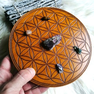 Peut inclure: Un plateau circulaire en bois avec un motif de fleur de vie gravé. Cinq cristaux sont placés sur le plateau : un œil de tigre, un cristal rose, un cristal violet, un cristal vert et un cristal transparent.