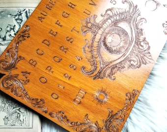 May include: A rectangular wooden spirit board featuring a laser-engraved design of a large central eye surrounded by ornate scrollwork. The board includes the alphabet, numbers 0-9, the words AU REVOIR, OUI, and NON, and a sun and moon symbol.