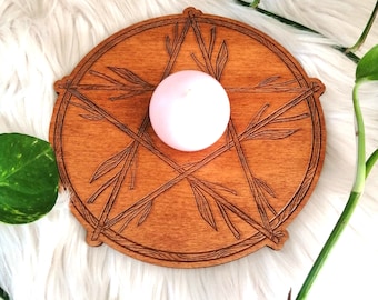 Puede incluir: Un círculo de madera con un diseño de estrella de cinco puntas y una vela rosa en el centro. La estrella está hecha de líneas finas que se asemejan a ramas.