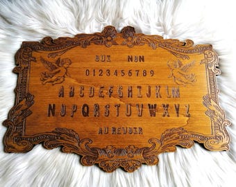 May include: Wooden Ouija board with a brown stain. The board features a decorative frame with cherub figures on each side. The board includes the alphabet, numbers 0-9, "Oui", "Non", and "Au Revoir".