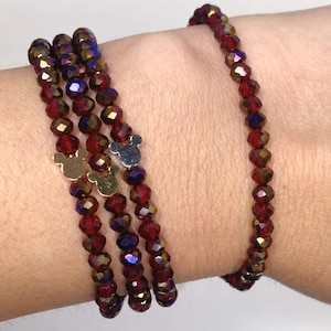 May include: A collection of red and gold beaded bracelets with Mickey Mouse charms. The bracelets are made of small, faceted beads that catch the light. The charms are gold and silver. The bracelets are stacked on a wrist.