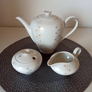 Könnte beinhalten: Ein weißes Porzellan-Tee-Set mit einem Blumenmuster. Das Set enthält eine Teekanne, eine Milchkännchen und eine Zuckerdose. Die Teekanne hat einen goldenen Rand um den Deckel und den Ausguss. Die Milchkännchen und die Zuckerdose haben einen goldenen Rand um den Deckel.