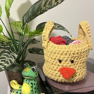 May include: A yellow crocheted basket with a cute face, featuring black eyes and a red nose. The basket is filled with colourful yarn balls and has two handles. A green frog figurine is sitting on a table next to the basket.