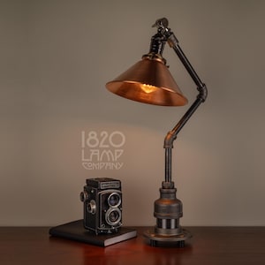 May include: A black and copper industrial style table lamp with a vintage camera on a black book on a wooden surface. The lamp has a copper shade and a black metal base. The text "1820 LAMP COMPANY" is visible on the wall behind the lamp.