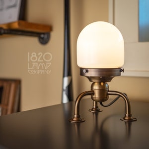 May include: A white glass globe lamp with a brass base and three legs. The lamp is on a dark gray surface. The text "1820 LAMP COMPANY" is visible in the background.
