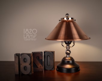May include: A copper-toned table lamp with a black base and a copper-colored shade. The lamp is turned on and the light is shining. The lamp is sitting on a wooden table with the words "1820 LAMP COMPANY" printed in black on a white background.