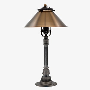 May include: A brass and black metal table lamp with a cone-shaped shade. The lamp has a steampunk aesthetic and is made of industrial pipes.