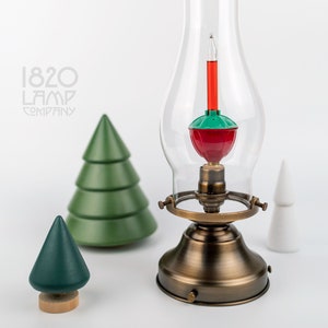 May include: A vintage-style lamp with a red and green Christmas light bulb inside a clear glass globe. The lamp has a bronze base and a handle. Two green wooden Christmas trees and a white cone-shaped decoration are also present. The logo for "1820 Lamp Company" is in the upper left.