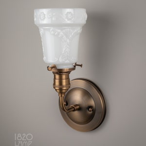 May include: A brass wall sconce with a white frosted glass shade featuring an embossed floral design. The sconce has a round back plate and a curved arm.