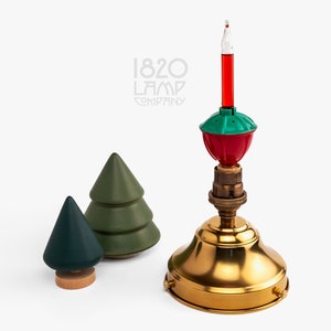 May include: A red and green Christmas light bulb with a gold base and a clear glass tube. The bulb is sitting on a gold metal base. The base is surrounded by two small green pine trees.