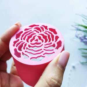 Pode incluir: Um objeto cilíndrico rosa brilhante com uma superfície superior plana apresentando um padrão floral detalhado cortado a laser. O design consiste em formas orgânicas concêntricas que lembram as pétalas de uma rosa ou peônia em flor.