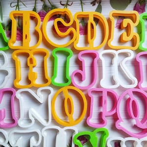 Puede incluir: Un juego de cortadores de galletas de plástico con forma de alfabeto en varios tonos de verde, amarillo, rosa y blanco. Los cortadores están dispuestos en orden alfabético de la A a la Q.