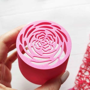 Puede incluir: Un objeto cilíndrico rosa con un diseño recortado en forma de rosa en la parte superior. El objeto se sostiene en una mano, con tela roja y encaje en el fondo. El diseño es un elemento decorativo.