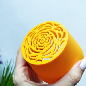 May include: A bright yellow cylindrical object with a floral design cut into the top. The object is held in a hand with a light blue background. The floral design is a series of curved lines that create a spiral pattern.