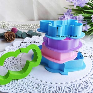 May include: A collection of plastic cookie cutters in various colours: blue, purple, pink, and green. The blue cutter is 15 mm (0.59 inches) tall, the green cutter is 6 mm (0.24 inches) wide, and the pink cutter is 1.5 mm (0.059 inches) tall.