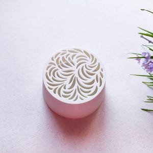 May include: A circular white object with a smooth, solid base and a top surface featuring an intricate, cut-out pattern of swirling, leaf-like shapes. The design consists of repeating curved lines that radiate from the center outward.