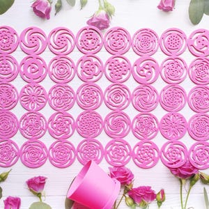 Könnte beinhalten: Eine Sammlung von pinkfarbenen, kreisförmigen Ausstechern in Blumenform mit verschiedenen, aufwendigen Designs. Die Ausstecher sind in Reihen auf einer weißen Oberfläche angeordnet, mit rosa Rosen und einem pinkfarbenen Behälter im Hintergrund. Ideal zum Backen und Basteln.