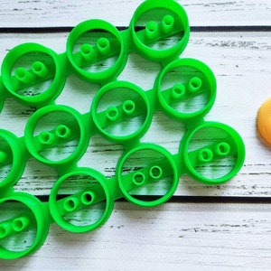 Può includere: Tagliabiscotti in plastica verde a forma di bottoni con un numero diverso di fori. Un solo bottone giallo con un viso accigliato è a destra dei tagliabiscotti.