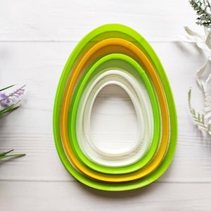 Puede incluir: Conjunto de cinco cortadores de galletas de plástico verde y amarillo con forma de huevo. Los cortadores están anidados uno dentro del otro, con el cortador más grande en el exterior y el más pequeño en el interior.