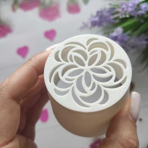 Puede incluir: Difusor cilíndrico blanco con un diseño floral recortado. La parte superior del difusor tiene un patrón detallado de pétalos superpuestos. El difusor se sostiene en una mano, con un fondo borroso de flores rosas y moradas.
