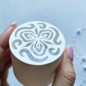 Könnte beinhalten: Ein zylindrisches weißes Objekt mit einer flachen Oberseite, die ein ausgeschnittenes Design einer stilisierten dreiblättrigen Blume umgeben von geschwungenen Mustern aufweist. Das Material hat eine matte, texturierte Oberfläche und ist hohl.