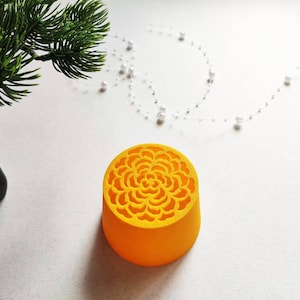 Könnte beinhalten: Ein leuchtend gelbes, zylindrisches Objekt mit einem detaillierten, filigranen Blumenmuster als Ausschnitt auf der Oberseite. Der Gegenstand hat eine glatte, feste Basis und eine flache, kreisförmige Oberseite mit einem sich wiederholenden, symmetrischen Blütenblattdesign.