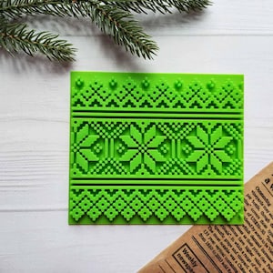 Puede incluir: Un molde de silicona verde brillante con un patrón geométrico detallado, con diseños de copos de nieve y diamantes pixelados. El molde es rectangular y probablemente se utiliza para manualidades o repostería.
