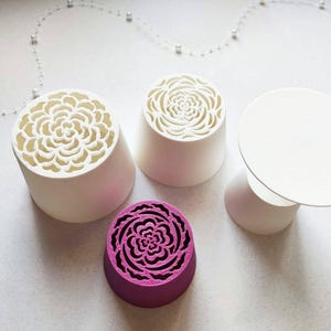 Pode incluir: Três objetos cilíndricos com bases cónicas e um topo circular sólido. Dois objetos brancos apresentam padrões florais intrincados cortados a laser no topo. Um objeto magenta também exibe um design floral correspondente no topo.