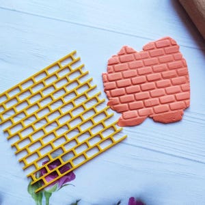 May include: A yellow brick-patterned stencil and a terracotta-colored clay brick impression are displayed on a light blue surface. A wooden rolling pin is visible in the background, suggesting a crafting or baking theme.