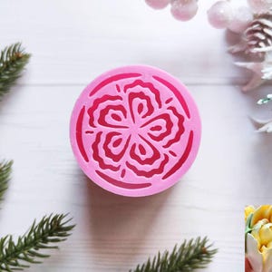 May include: A pink, circular silicone mold with a floral design. The mold features a detailed, symmetrical pattern of layered petals. The image is set against a white background with sprigs of evergreen and frosted winter foliage.