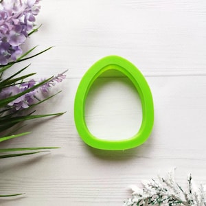 Puede incluir: Un cortador de galletas de plástico verde brillante con forma de huevo. El cortador está sobre una superficie de madera blanca.