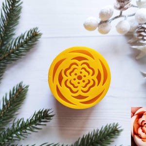 May include: A bright yellow, circular mold with a floral design. The mold is surrounded by evergreen branches and decorative winter foliage. The mold's design is a detailed flower pattern.