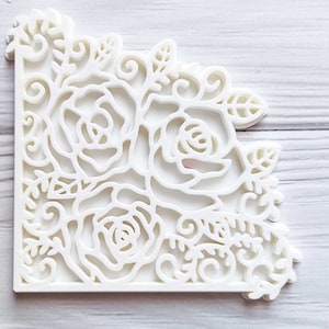 May include: A white, corner-shaped decorative piece featuring a floral design. The piece is cut out with intricate details of roses, leaves, and swirling patterns. The design is symmetrical and appears to be made of a solid, opaque material.