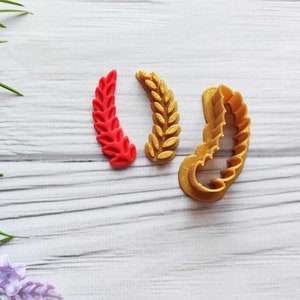 May include: Three gold-coloured cookie cutters shaped like wheat stalks. The cutters are on a white wooden surface.